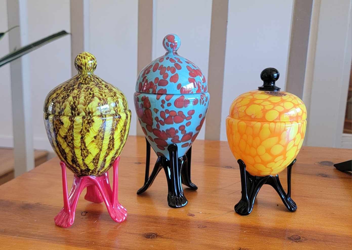 Three colorful ceramic jars with lids, each on three-legged stands, placed on a wooden table.